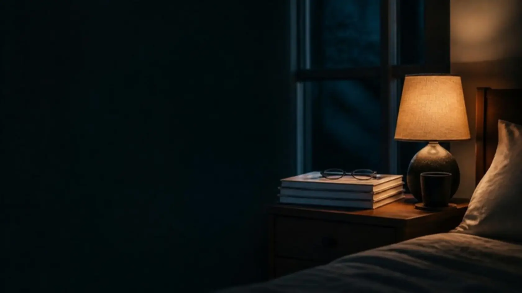 A bedroom at night, lit only by a bedside lamp, with a stack of research journals and reading glasses